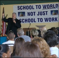 Audience listening to speaker with sign in background: school to world, not just school to work